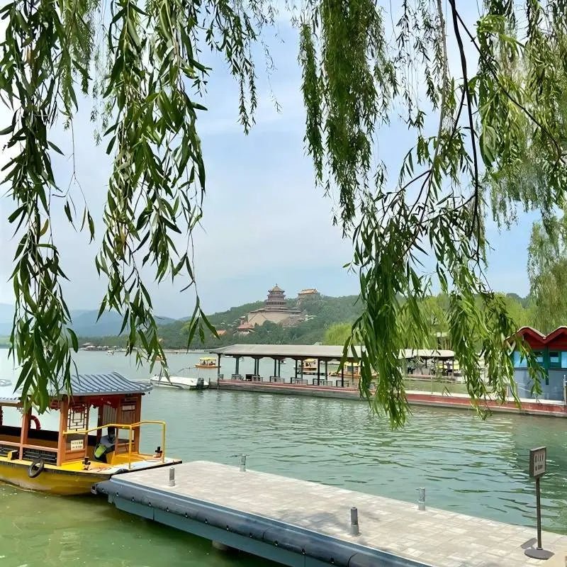 Weeping willow branches framing Kunming Lake at the Summer Palace in Beijing, traditional yellow Chinese tour boat docked at the pier with Tower of Buddhist Incense in the distance